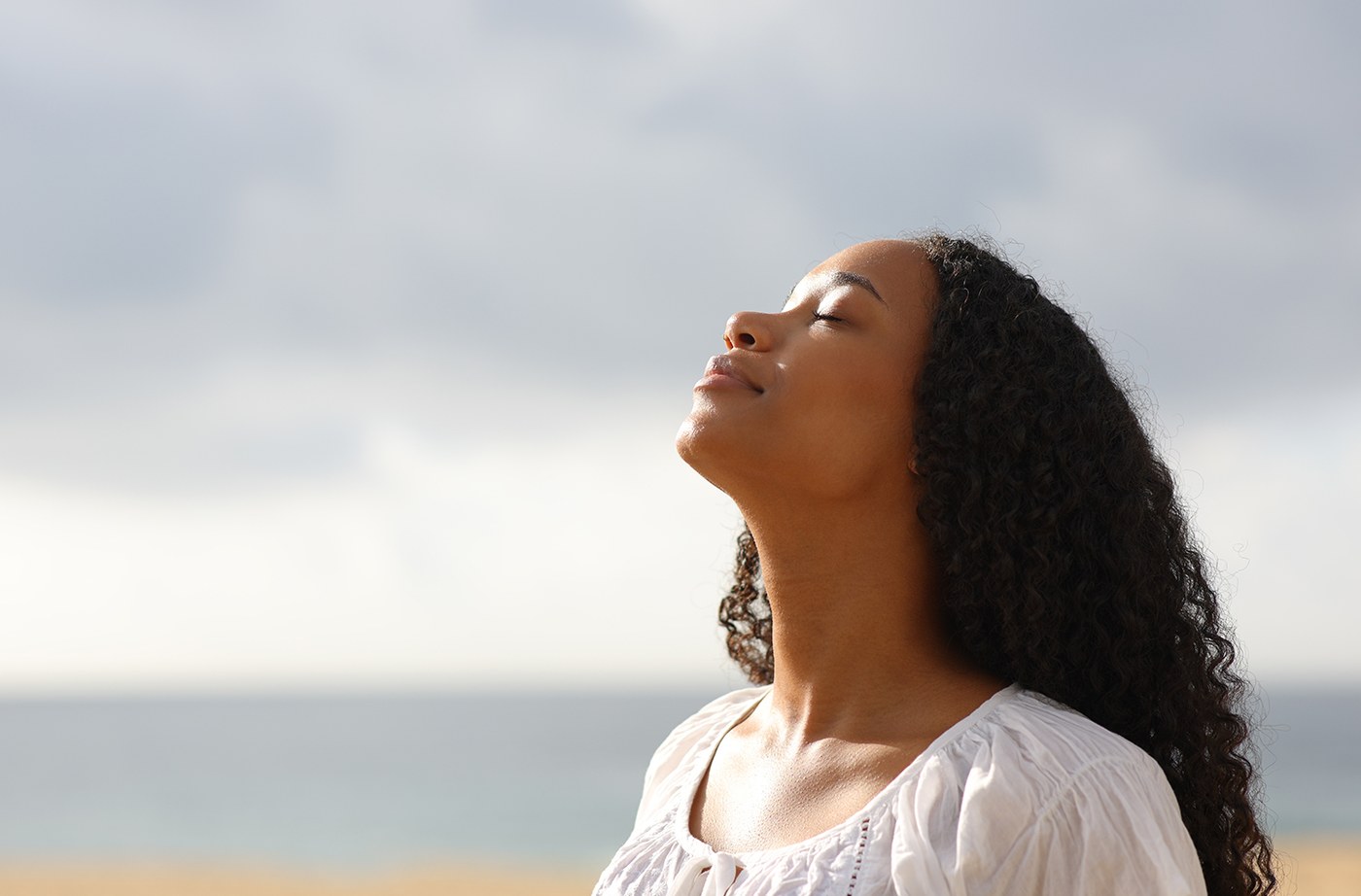 A woman standing outdoors near the ocean with her eyes closed, taking a slow breath